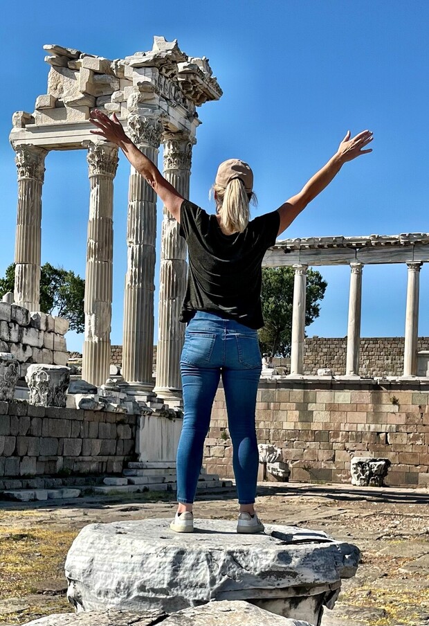 A person stands on a stone with arms raised, facing ancient ruins featuring columns under a clear blue sky.